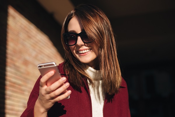 Young smiling Ukrainian lady in sunglasses standing on the street and happily using her mobile phone